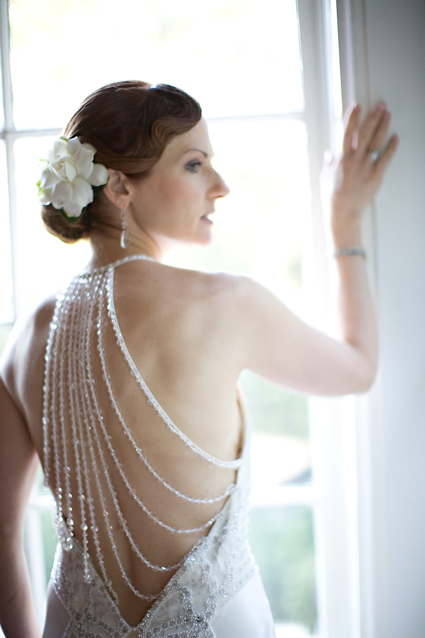 Beautiful bride looking out window wearing a gorgeous white backless wedding gown with embellishments and a white floral hair piece - wedding photo by Meg Perotti